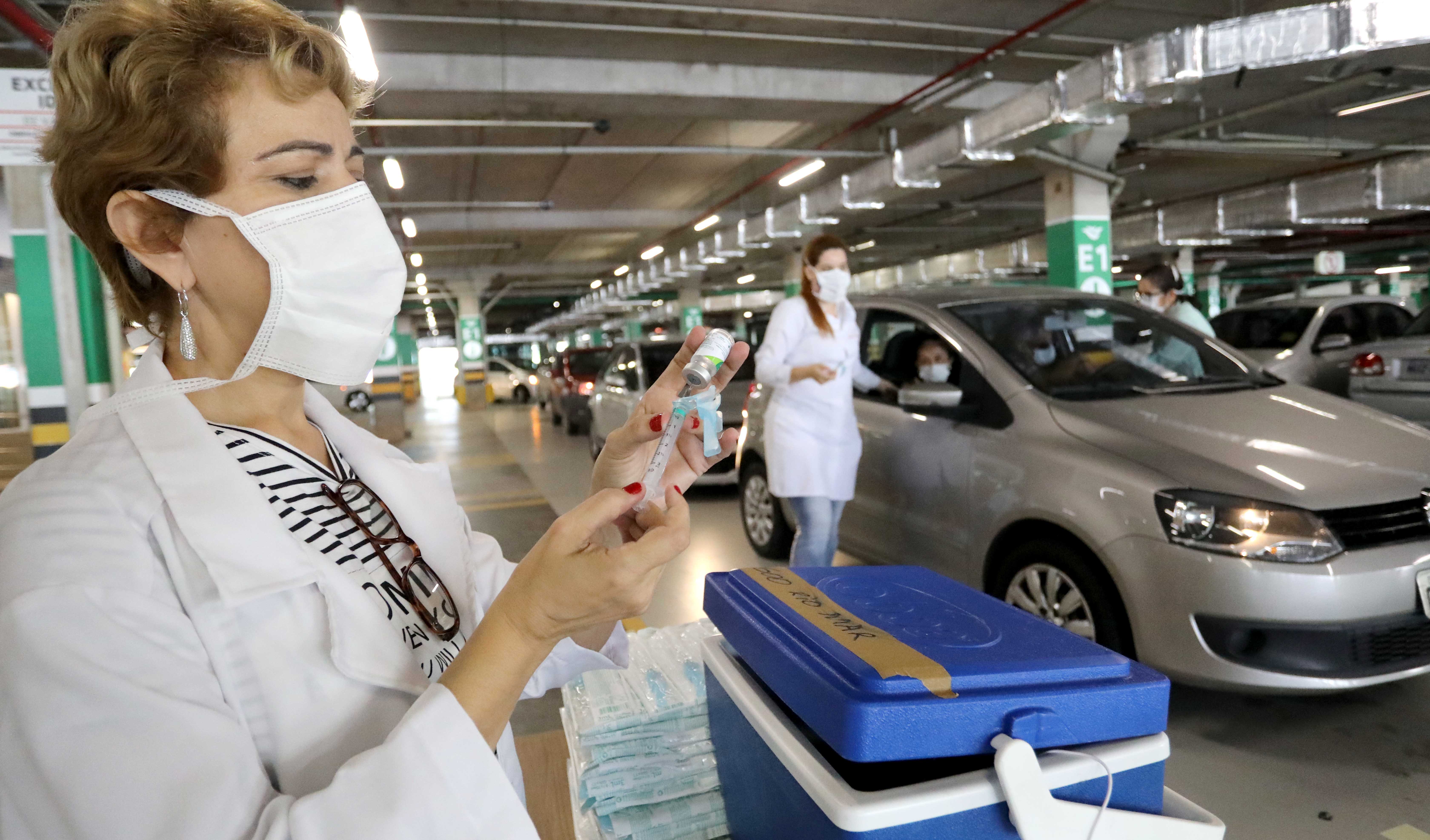 enfermeira manuseando seringa e carro ao fundo em estacionamento de shopping